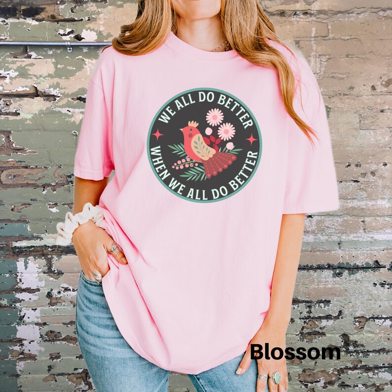 Puede incluir: Camiseta rosa con un gr&aacute;fico de un p&aacute;jaro rojo con flores y el texto "We all do better when we all do better".