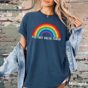 May include: A dark blue t-shirt with a rainbow graphic and the text "FOXTROT DELTA TANGO" printed on the front. The shirt is being worn by a person with long blonde hair.