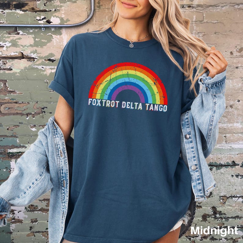 May include: A dark blue t-shirt with a rainbow graphic and the text "FOXTROT DELTA TANGO" printed on the front. The shirt is being worn by a person with long blonde hair.