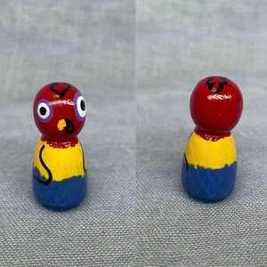 May include: A hand-painted wooden doll with a red head, yellow body, and blue legs. The doll has a black heart shape painted on its head and two large blue eyes.