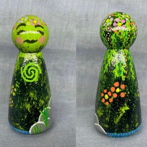 May include: A green wooden doll with a painted face and a green swirl design on its chest. The doll is decorated with green, orange, and pink paint, resembling a forest scene.