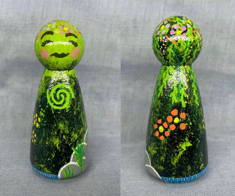 May include: A green wooden doll with a painted face and a green swirl design on its chest. The doll is decorated with green, orange, and pink paint, resembling a forest scene.