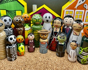 May include: A collection of hand-painted wooden Halloween peg dolls depicting classic monsters and spooky characters. The figures include a mummy, skeleton, werewolf, vampire, creature, ghost, and pumpkin-headed figures. The background features painted houses.