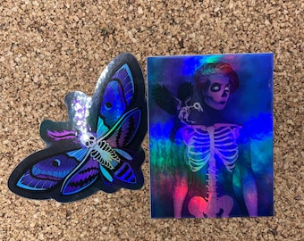 May include: Two iridescent stickers. One features a moth with blue, purple, and black wings. The other features a skeleton with a bird perched on its shoulder. Both stickers have a holographic finish.