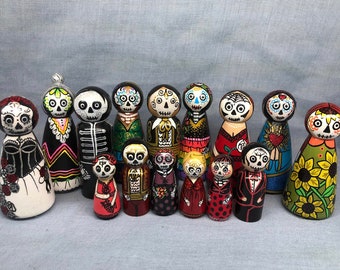 Puede incluir: Un conjunto de 15 muñecas de madera pintadas a mano vestidas con coloridos atuendos del Día de Muertos. Las muñecas presentan rostros esqueléticos en blanco y negro con coloridos patrones florales y geométricos.