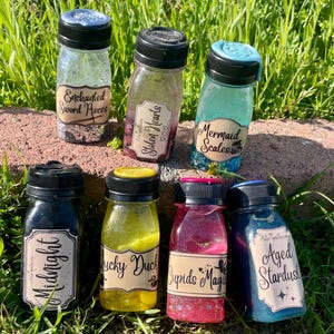 Puede incluir: Siete pequeñas botellas de vidrio con tapas negras llenas de purpurina y líquidos de colores. Cada botella tiene una etiqueta con un nombre y un tema diferente, como "Medianoche", "Piezas de espada encantadas", "Corazones robados", "Escamas de sirena", "Pato de la suerte", "Magia de Cupido" y "Polvo de estrellas envejecido".