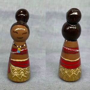 May include: Hand-painted wooden doll with brown hair, a red and brown dress, and a gold and brown skirt. The doll is wearing a necklace with a blue and gold pendant.