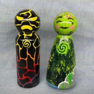 May include: Two hand-painted wooden peg dolls, one with a black and yellow lava-like design and the other with a green and black design with a swirl pattern.