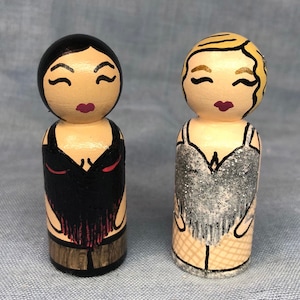 Puede incluir: Dos muñecas de madera pintadas a mano vestidas con atuendos glamorosos. Una muñeca tiene el pelo negro y lleva un vestido negro con detalles rojos. La otra muñeca tiene el pelo rubio y lleva un vestido de lentejuelas plateadas.