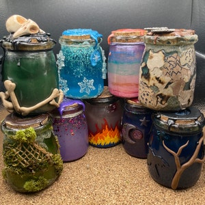 May include: A collection of 10 small glass jars with cork lids. The jars are filled with colorful wax and have various designs, including snowflakes, flames, and a tree branch.