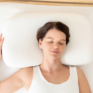 May include: A woman with brown hair is sleeping on a white pillow with her eyes closed. She is wearing a white tank top.