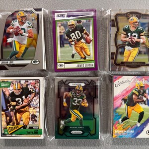 May include: Six football trading cards featuring Green Bay Packers players. The cards are in protective sleeves. Players include Jordan Love, James Lofton, Aaron Rodgers, Jim Taylor, Aaron Jones, and Josh Jacobs. The cards have various designs and backgrounds.