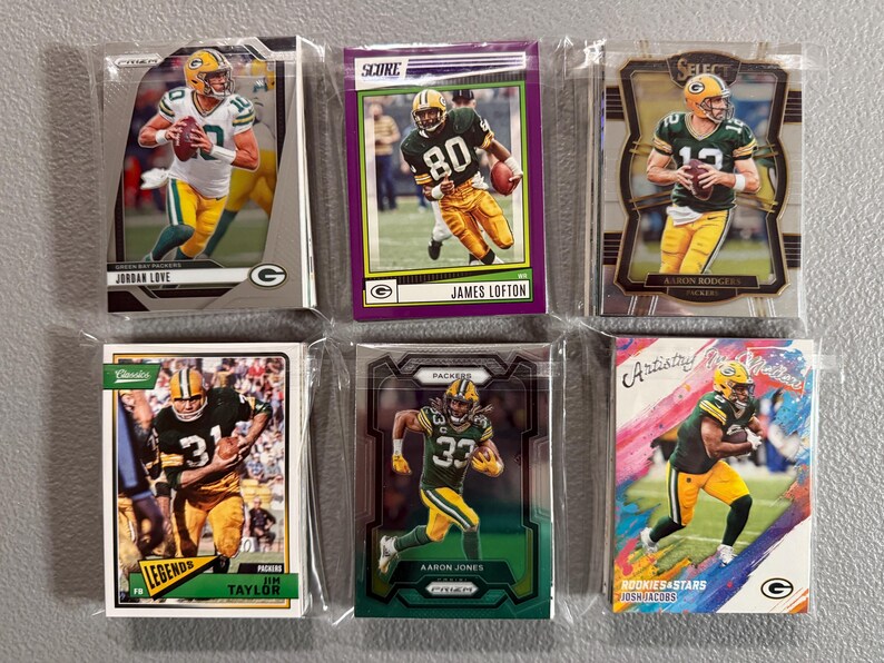 May include: Six football trading cards featuring Green Bay Packers players. The cards are in protective sleeves. Players include Jordan Love, James Lofton, Aaron Rodgers, Jim Taylor, Aaron Jones, and Josh Jacobs. The cards have various designs and backgrounds.