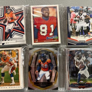 May include: Six Denver Broncos football trading cards in protective sleeves. The cards feature players like Shannon Sharpe, Von Miller, and Demaryius Thomas. Some cards have the text "ROOKIES & STARS" and "PLAYOFF".