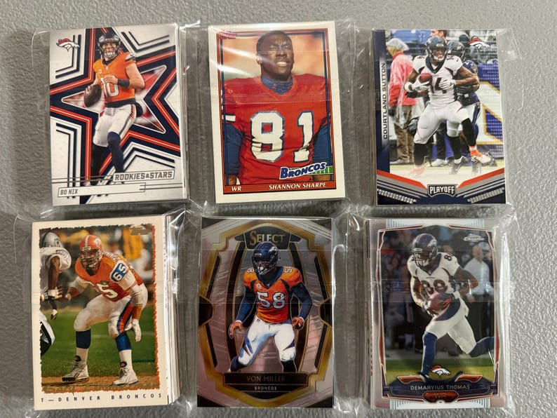 May include: Six Denver Broncos football trading cards in protective sleeves. The cards feature players like Shannon Sharpe, Von Miller, and Demaryius Thomas. Some cards have the text "ROOKIES & STARS" and "PLAYOFF".