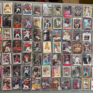 May include: A collection of numerous baseball trading cards arranged in a grid, each encased in a clear plastic protective sleeve. The cards feature various players in action poses, portraits, and team uniforms with colorful graphic designs and text.
