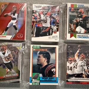 May include: A collection of six football trading cards featuring Cincinnati Bengals players. The cards are in protective sleeves and showcase players like Joe Burrow, Ja'Marr Chase, and Chad Johnson. The cards have various designs and are from different sets.