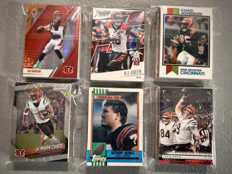 May include: A collection of six football trading cards featuring Cincinnati Bengals players. The cards are in protective sleeves and showcase players like Joe Burrow, Ja'Marr Chase, and Chad Johnson. The cards have various designs and are from different sets.