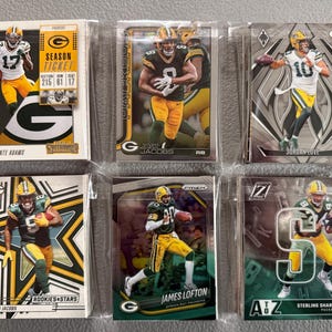 May include: A collection of six football trading cards featuring players in green and yellow uniforms. The cards are in protective sleeves. Players include Davante Adams, Josh Jacobs, James Lofton, and Sterling Sharpe.