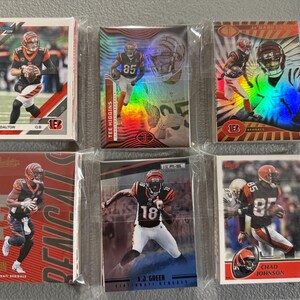 May include: A collection of six Cincinnati Bengals football trading cards, each encased in a protective sleeve. The cards feature images of players like Andy Dalton, Tee Higgins, and Chad Johnson, with team colors of black, orange, and white.