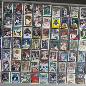 May include: A collection of numerous rectangular baseball trading cards featuring various players in action poses, batting, pitching, or fielding. Each card is individually encased in a clear plastic protective sleeve, arranged in a grid pattern.