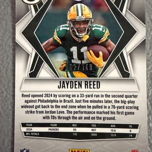 May include: A football trading card featuring Jayden Reed of the Packers. The card has a photo of Reed in his green and gold uniform, holding a football. The card includes statistics and the text "2025 Panini - Phoenix Football."
