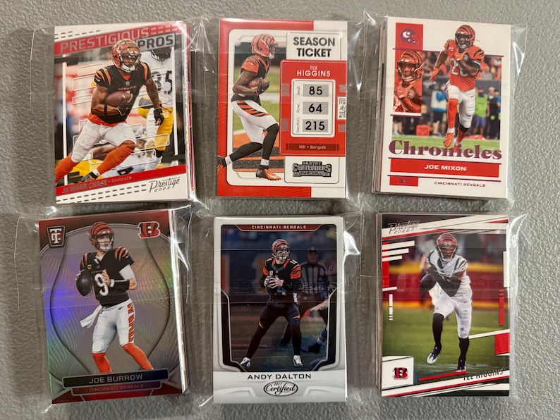 May include: Six trading cards featuring Cincinnati Bengals players, including Joe Burrow, Tee Higgins, and Joe Mixon. The cards are in protective sleeves and display action shots of the players in their uniforms. The cards have the words "Prestige," "Chronicles," and "Certified."