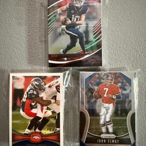 May include: Three Denver Broncos football trading cards in protective sleeves. The cards feature players in action, including a card with the text "John Elway" and another with "Von Miller-LB". The cards are in various designs and colors.