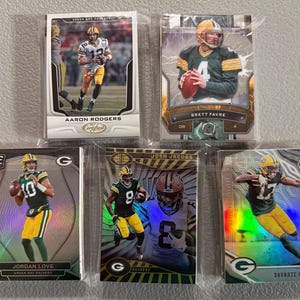 May include: Five Green Bay Packers football trading cards featuring Aaron Rodgers, Brett Favre, Jordan Love, Josh Jacobs, and Davante Adams. The cards are in protective sleeves and feature action shots of the players in their uniforms.