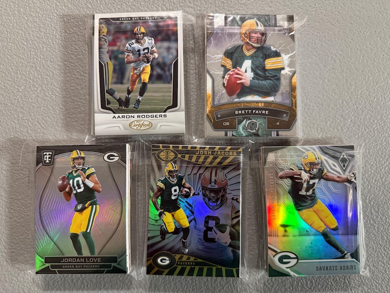 May include: Five Green Bay Packers football trading cards featuring Aaron Rodgers, Brett Favre, Jordan Love, Josh Jacobs, and Davante Adams. The cards are in protective sleeves and feature action shots of the players in their uniforms.