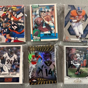 May include: A collection of six Denver Broncos football trading cards in protective sleeves. The cards feature players like John Elway, Tony Dorsett, Demarcus Ware, Courtland Sutton, and Demaryius Thomas. The cards have various designs and logos.