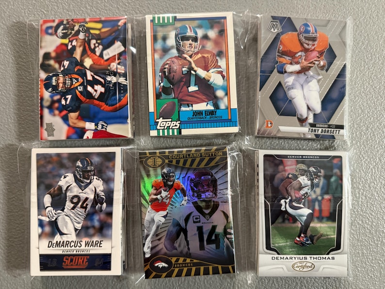 May include: A collection of six Denver Broncos football trading cards in protective sleeves. The cards feature players like John Elway, Tony Dorsett, Demarcus Ware, Courtland Sutton, and Demaryius Thomas. The cards have various designs and logos.