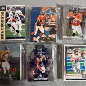 May include: A collection of six Denver Broncos football trading cards in protective sleeves. The cards feature various players, including Peyton Manning and Tim Tebow. The cards are from different sets, with the "NFL Legends" and "Prizm" logos visible.