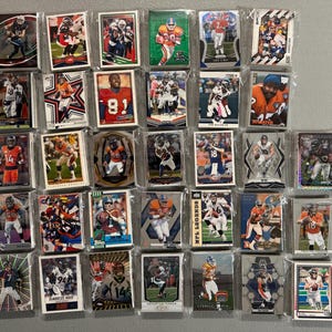 May include: A collection of football trading cards, each encased in a protective sleeve. The cards feature various players in action, with team colors and logos prominently displayed. The cards are arranged on a neutral gray surface.