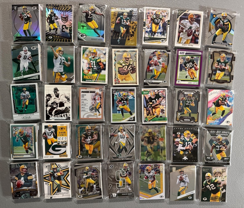 May include: A collection of Green Bay Packers football trading cards, featuring players in their green and gold uniforms. The cards are in protective sleeves, showcasing various action shots and player portraits. Some cards have the Packers logo.