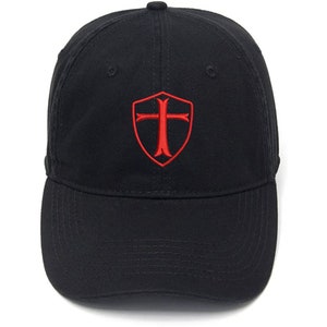 Puede incluir: Una gorra de béisbol negra con un escudo y una cruz bordados en rojo.