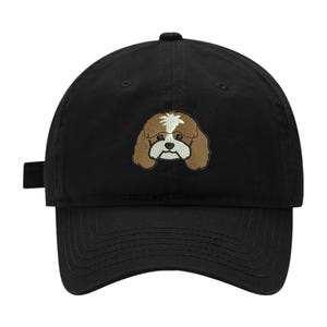 May include: A black baseball cap featuring an embroidered graphic of a brown and white dog face centered on the front panel. The cap has a curved brim, a structured crown, and a fabric strap with a metal buckle closure visible on the left side.