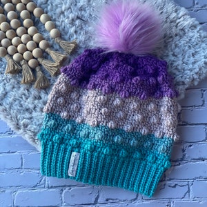 May include: A purple, beige, and teal crocheted beanie with a fluffy purple pom pom. The beanie has a label that reads "Daber Autumn Co."
