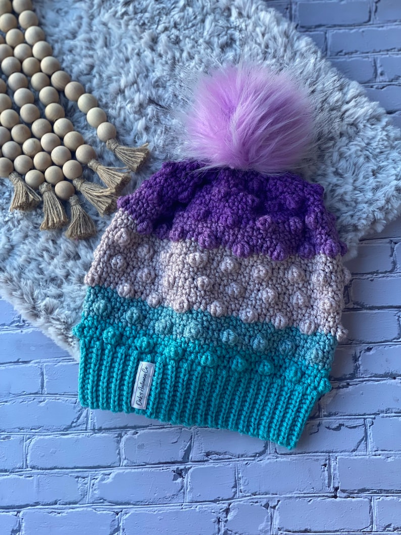 May include: A purple, beige, and teal crocheted beanie with a fluffy purple pom pom. The beanie has a label that reads "Daber Autumn Co."