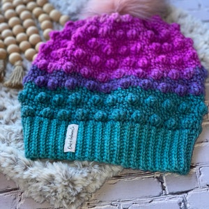 May include: A teal, purple, and pink crocheted beanie with a fluffy pom pom on top. The beanie has a textured, bumpy design and a label that reads "D&K Handmade Co."