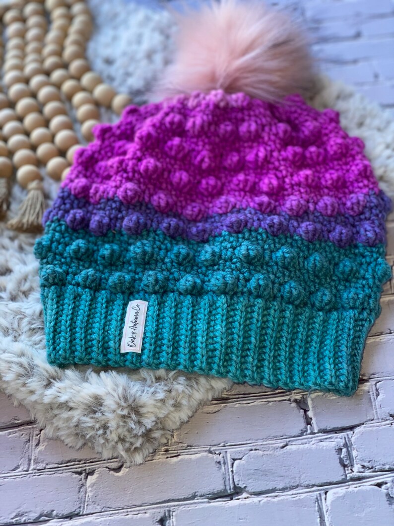 May include: A teal, purple, and pink crocheted beanie with a fluffy pom pom on top. The beanie has a textured, bumpy design and a label that reads "D&K Handmade Co."