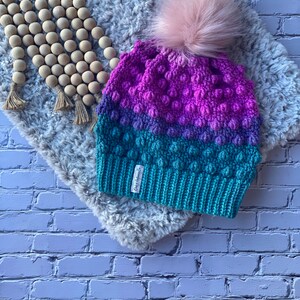 May include: A crocheted beanie with a pink pom pom. The beanie is made in a gradient of pink, purple, and teal colors. The beanie has a textured stitch pattern and a small tag that says "Cozy Knits".