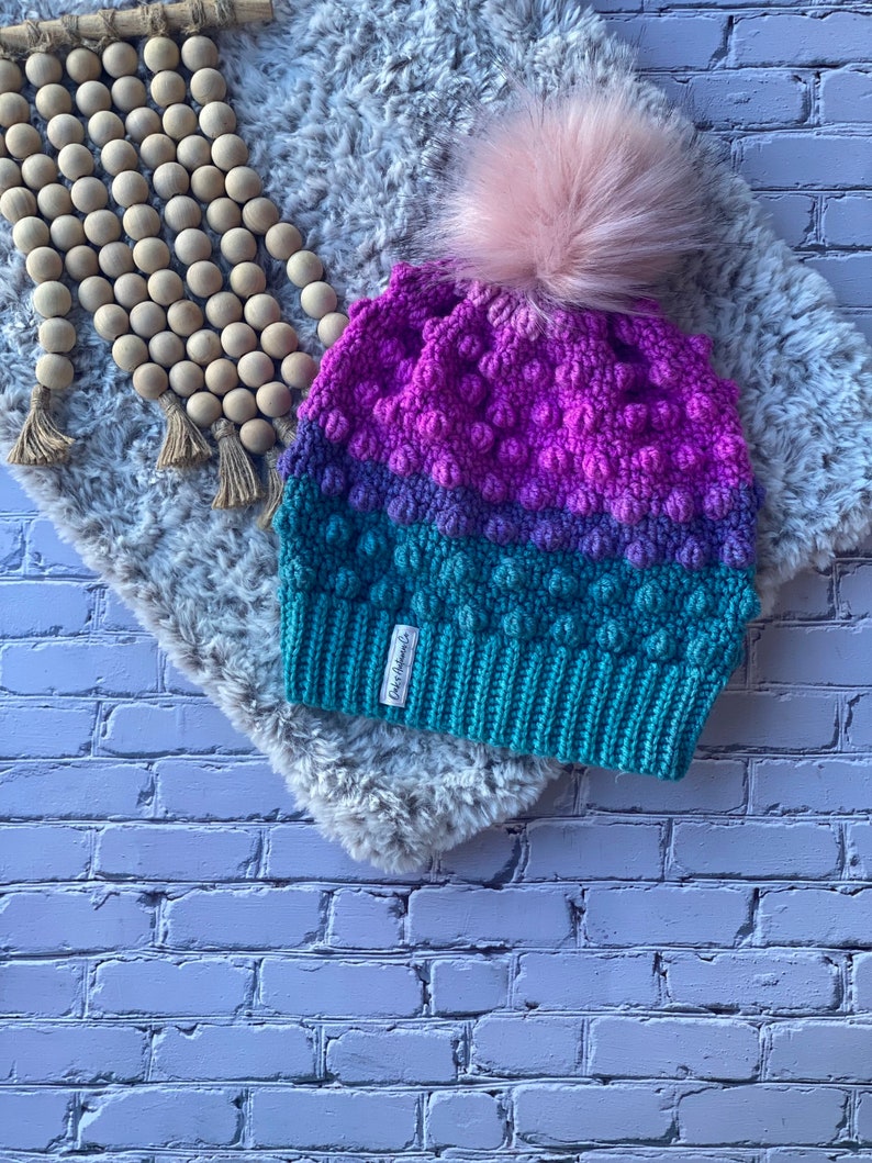 May include: A crocheted beanie with a pink pom pom. The beanie is made in a gradient of pink, purple, and teal colors. The beanie has a textured stitch pattern and a small tag that says "Cozy Knits".