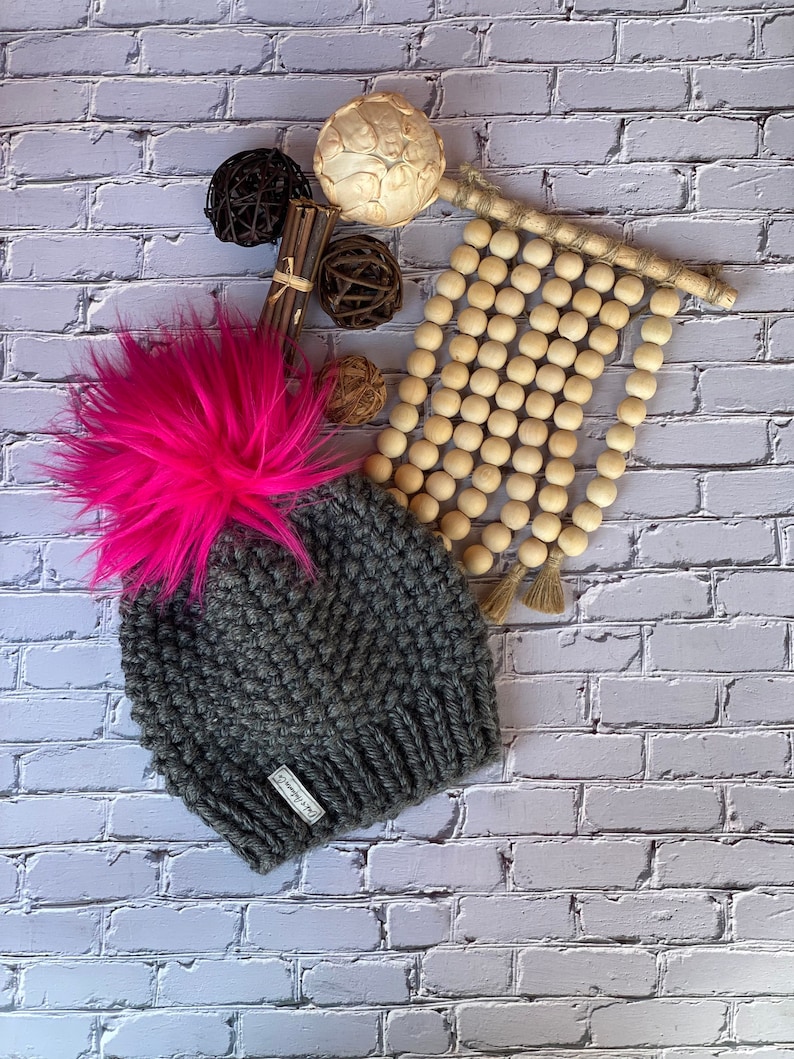 May include: A gray knitted beanie with a large pink pom pom on top. The beanie has a small white tag with the text "The Cozy Co." on it.