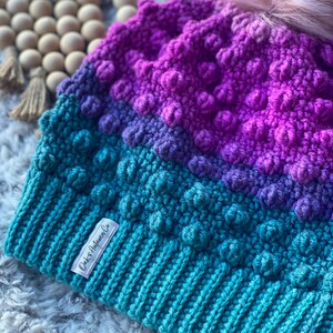 May include: A teal, purple, and pink crocheted beanie with a fluffy pom pom on top. The beanie has a textured, bobbly design and a white label that says "Oak & Autumn Co."