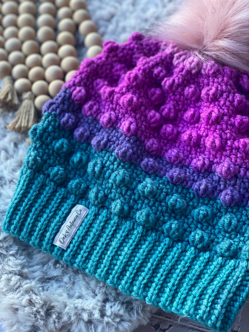 May include: A teal, purple, and pink crocheted beanie with a fluffy pom pom on top. The beanie has a textured, bobbly design and a white label that says "Oak & Autumn Co."
