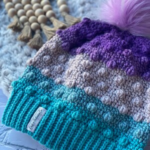 May include: A teal, gray, and purple crocheted beanie with a fluffy purple pom pom. The beanie has a textured design and a label that reads "Oaks Autumn Co."