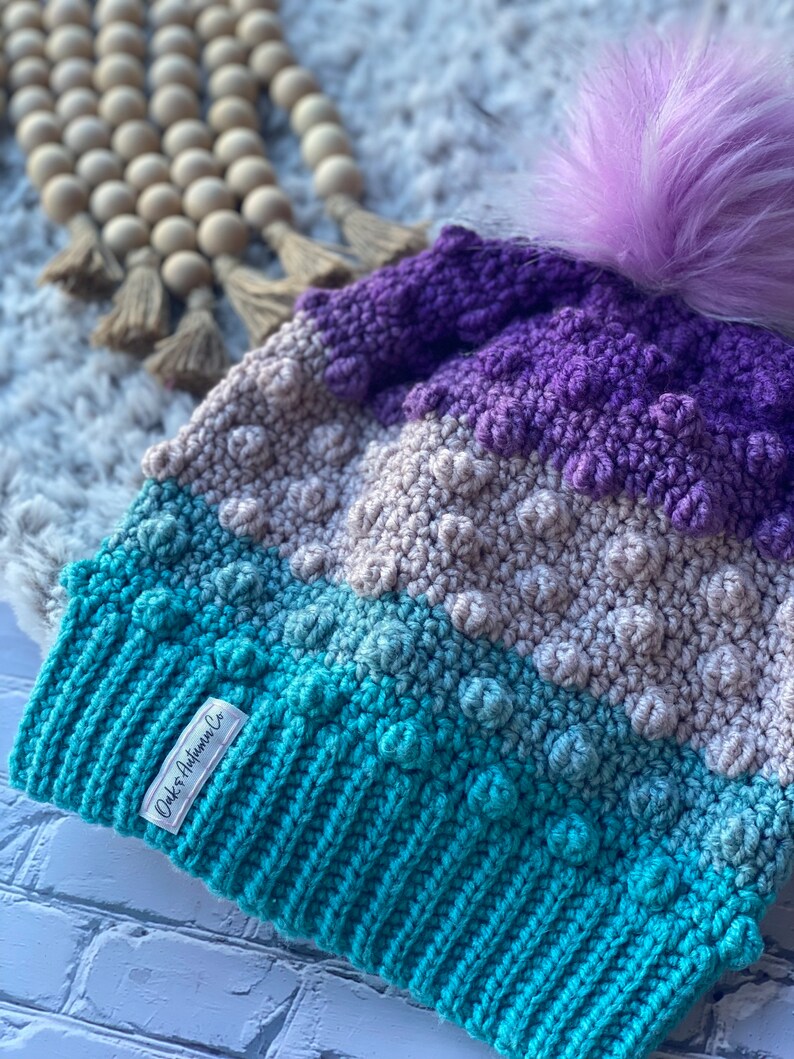 May include: A teal, gray, and purple crocheted beanie with a fluffy purple pom pom. The beanie has a textured design and a label that reads "Oaks Autumn Co."