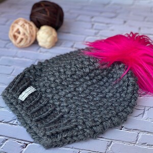 May include: A gray knitted beanie with a pink faux fur pom pom. The beanie has a small white label that says "Oaks Avenue Co."