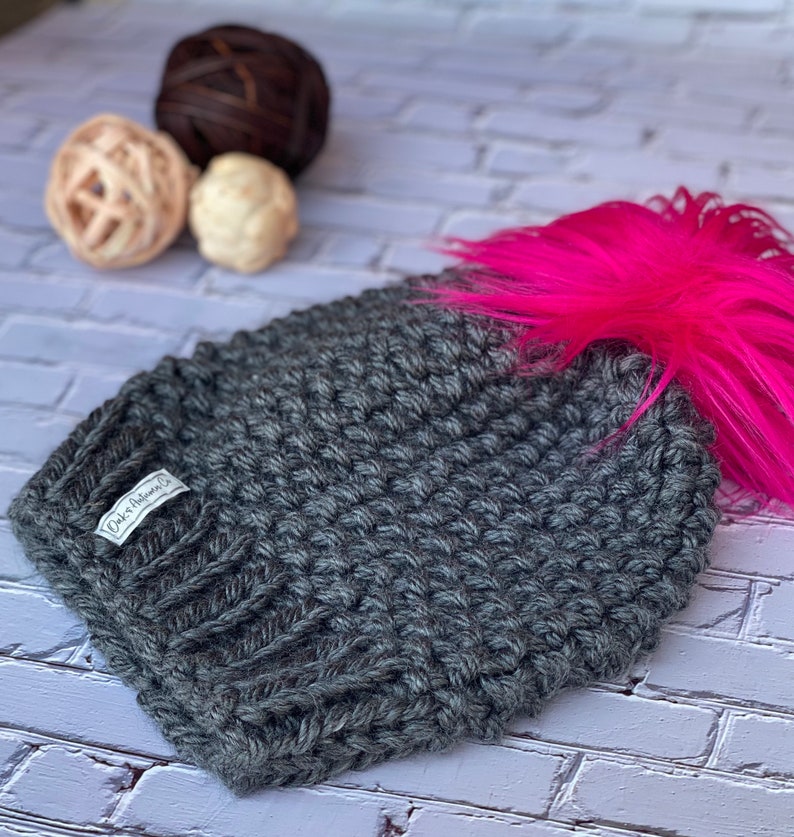 May include: A gray knitted beanie with a pink faux fur pom pom. The beanie has a small white label that says "Oaks Avenue Co."